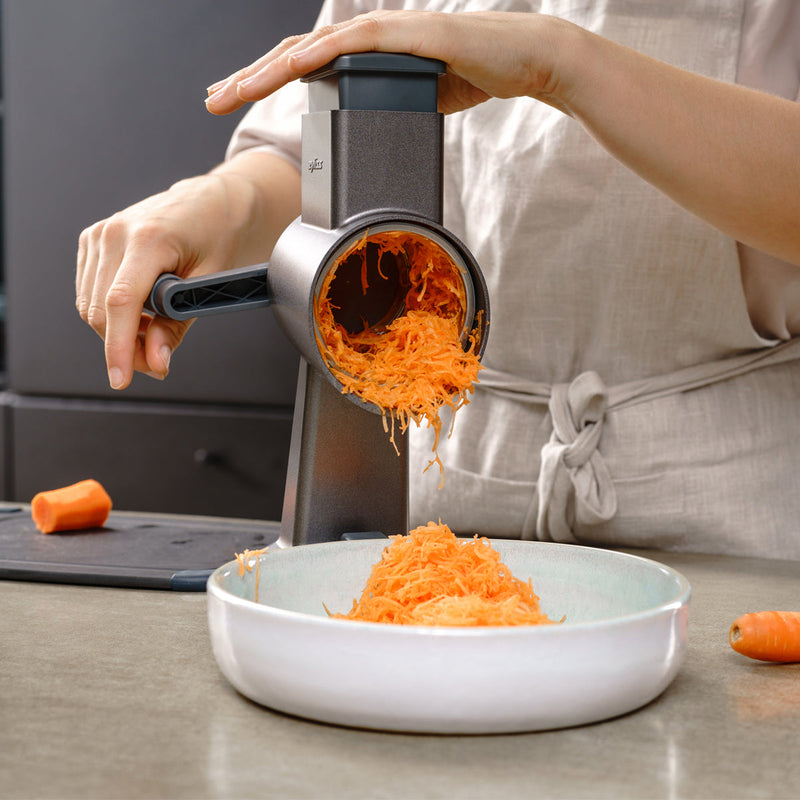 A person uses a rotary grater to shred carrots into a white bowl on a kitchen counter. A cutting board with a carrot is visible in the background.