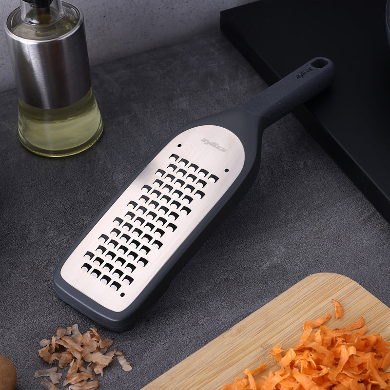 The Zyliss Coarse Grater, featuring a metal and plastic build with an ultra-sharp blade and black handle, sits on a gray countertop beside a cutting board with shredded carrots—an essential meal prep tool.
