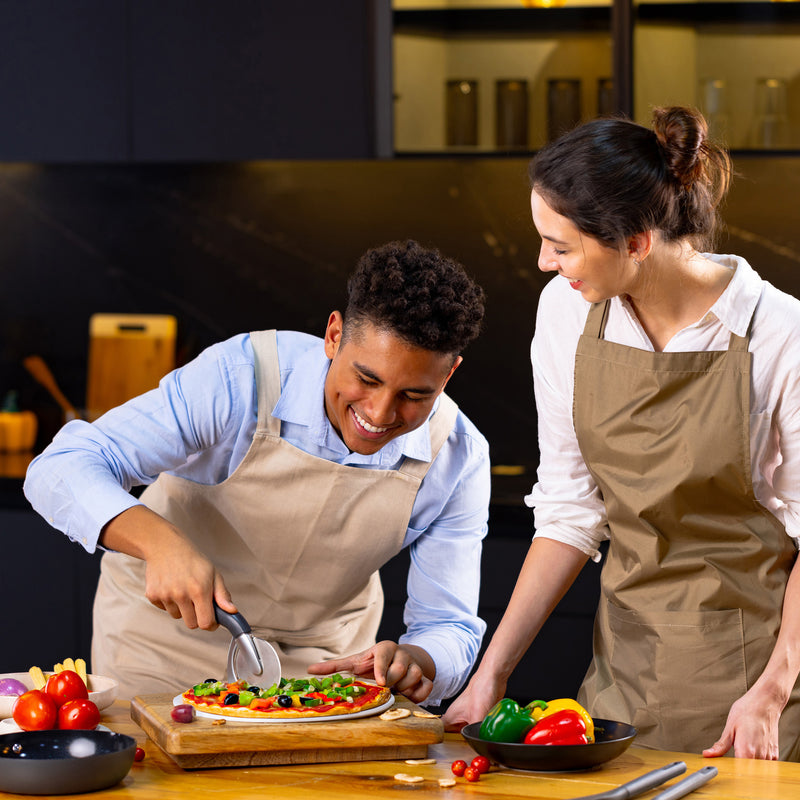 In a modern kitchen, two people in aprons smile while one slices a fresh pizza using the Zyliss Pizza & Pastry Cutter with an ergonomic handle. Colorful vegetables sit on the table, and they look like theyre enjoying cooking together.