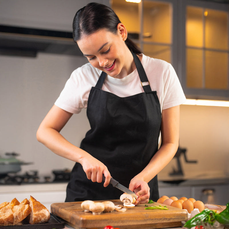 A woman in a black apron and white shirt smiles as she uses the Zyliss Vegetable Knife 9cm / 3½ with an eco-friendly handle to precisely slice mushrooms on a wooden board in her kitchen, surrounded by eggs, herbs, and bread.