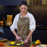 A woman in a beige apron uses the Zyliss Offset Knife 10cm/4 with an eco-friendly handle to slice a lemon on a cutting board in a modern kitchen, surrounded by fresh peppers and tomatoes, highlighting precise cutting.