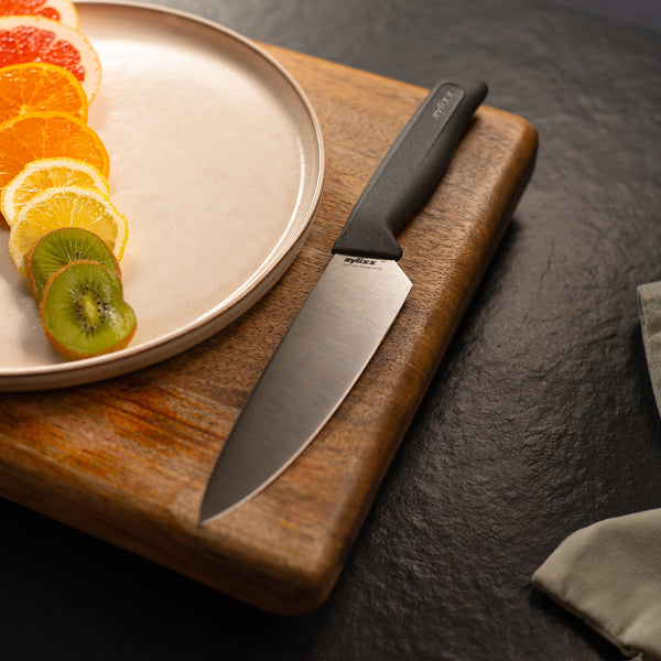 The Zyliss Chef Knife 15cm / 6 with an ergonomic handle lies on a wooden cutting board beside a plate of sliced citrus and kiwi, all arranged on a dark countertop.