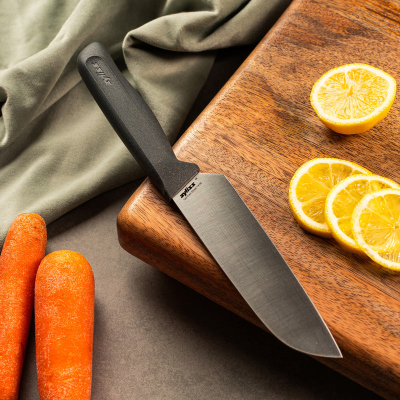 A Zyliss Santoku Knife 15cm / 6 with an ergonomic handle lies on a wooden cutting board beside sliced lemons, whole carrots, and a green cloth—all arranged on a gray surface for an inviting kitchen scene.