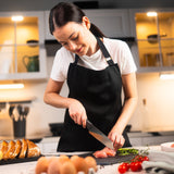 In a modern kitchen, a woman in a black apron slices raw chicken on a cutting board using the Zyliss Santoku Knife 15cm / 6 with an ergonomic handle. Bread, eggs, tomatoes, and herbs are arranged on the counter nearby.