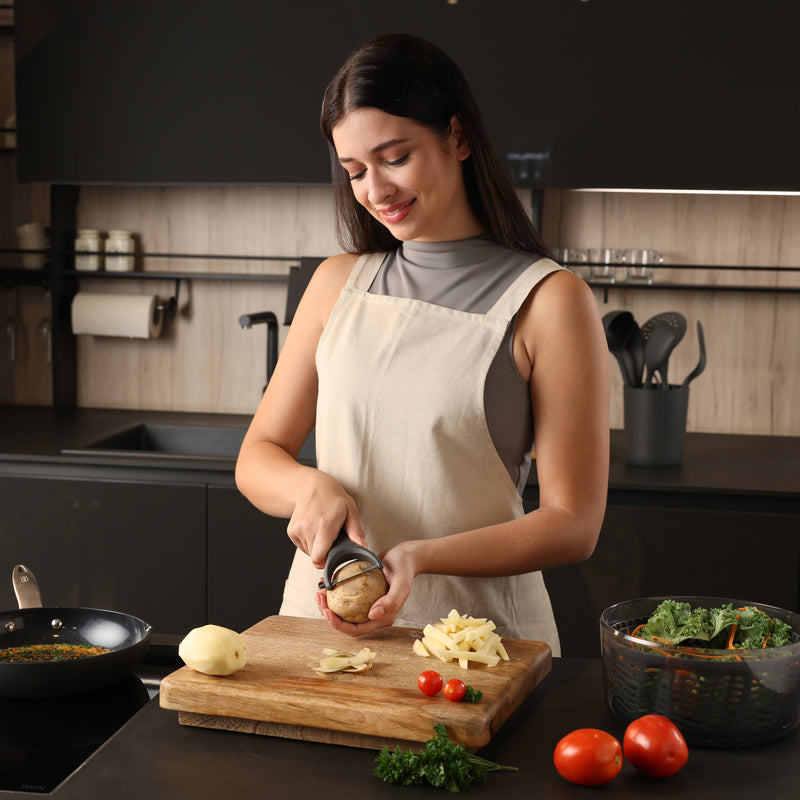 A woman in a light apron peels a potato at a kitchen counter, surrounded by chopped vegetables, tomatoes, a frying pan, and a bowl of greens. She is smiling and focused on her task.