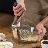 A person uses the Zyliss Potato Masher with a stainless steel head to mash cooked chickpeas in a glass bowl on a wooden surface, surrounded by garlic and small ingredient bowls.