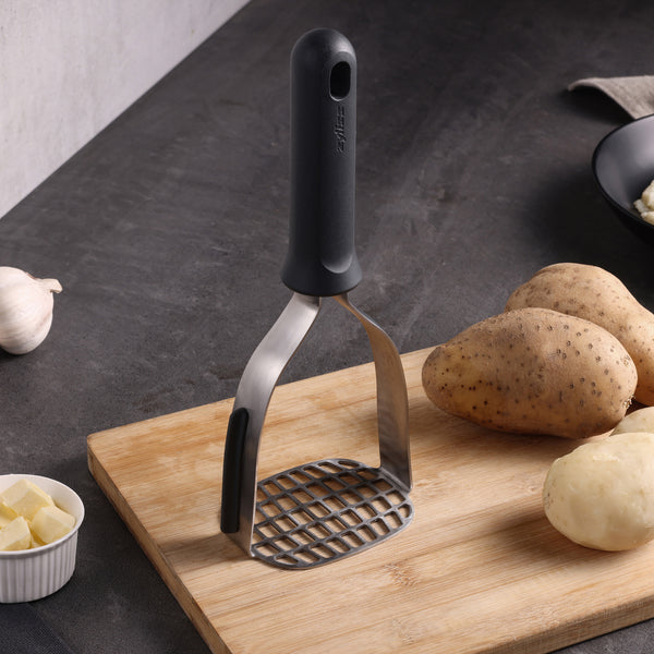 The Zyliss Potato Masher with an eco-friendly handle stands upright on a wooden cutting board beside raw potatoes, garlic, and butter cubes on a kitchen counter.