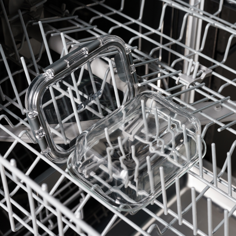 Two clear glass food containers with lids are placed inside the top rack of a dishwasher, ready to be washed. The dishwasher racks and prongs are visible around the containers.