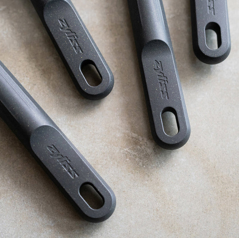 Four black kitchen utensil handles with oval holes at the ends are arranged diagonally on a light-colored surface. The handles are branded with the Zyliss logo.